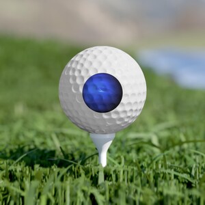 May include: A white golf ball with a dimpled surface texture sits atop a white golf tee. The center of the golf ball features a circular, glossy, deep blue graphic element that contrasts with the white surface of the ball.