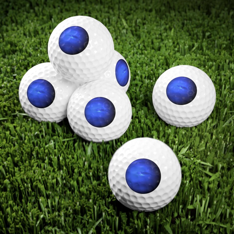 May include: A collection of five white golf balls, each featuring a prominent, circular, deep blue graphic in the center. The golf balls have a textured dimpled surface and are arranged in a cluster on a patch of green grass.
