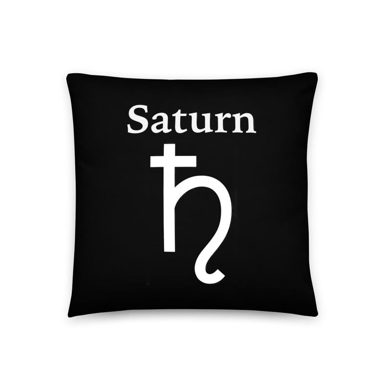 May include: A square black throw cushion featuring the white text Saturn centred above the white astronomical symbol for the planet Saturn. The design is printed clearly against the solid black fabric of the cushion cover.