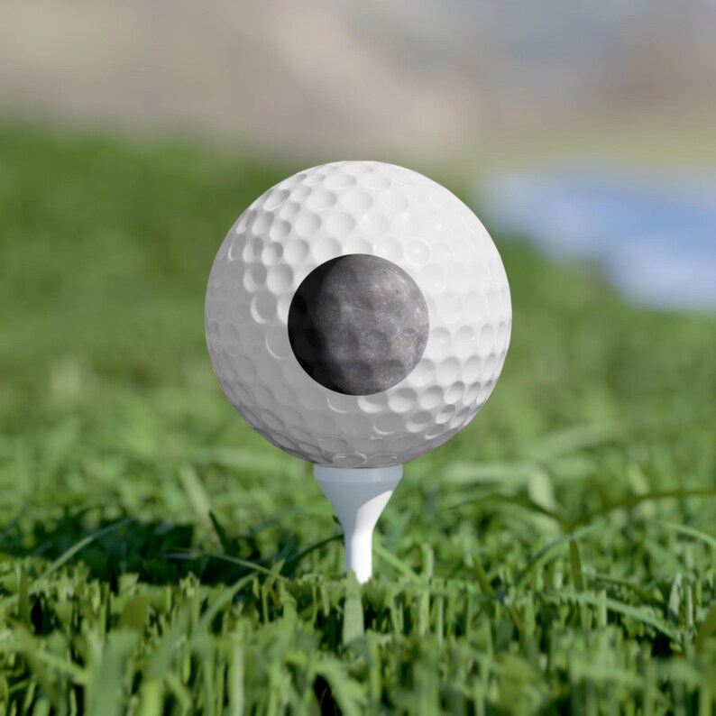 May include: A white golf ball with a dimpled surface resting on a white tee. The center of the ball features a circular graphic depicting a detailed, grey, cratered moon surface against the white background of the golf ball.
