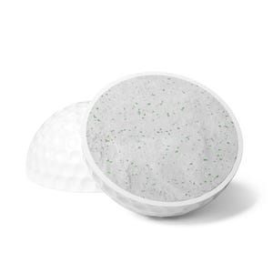 May include: A white golf ball cut in half, revealing a textured interior surface that is light gray with small, scattered green flecks. The exterior of the ball features the standard dimpled pattern characteristic of golf equipment.