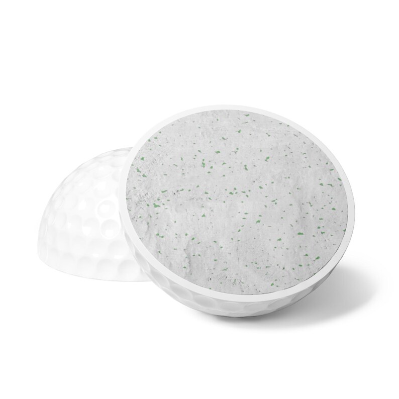 May include: A white golf ball cut in half, revealing a textured interior surface that is light gray with small, scattered green flecks. The exterior of the ball features the standard dimpled pattern characteristic of golf equipment.