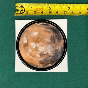 Puede incluir: Una pegatina circular con una imagen detallada del planeta Marte con texturas de superficie en tonos naranja, marr&oacute;n y blanco, rodeada por un borde negro fino. Una cinta m&eacute;trica amarilla muestra un di&aacute;metro de 7,6 cm.