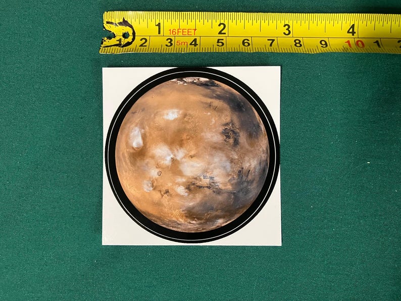 Puede incluir: Una pegatina circular con una imagen detallada del planeta Marte con texturas de superficie en tonos naranja, marr&oacute;n y blanco, rodeada por un borde negro fino. Una cinta m&eacute;trica amarilla muestra un di&aacute;metro de 7,6 cm.
