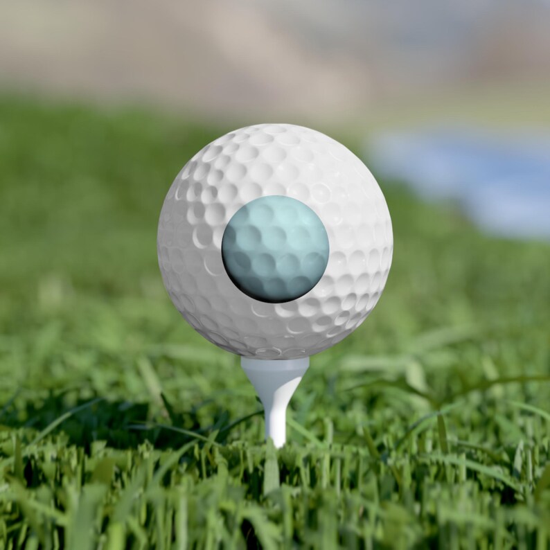 May include: A white golf ball with a dimpled surface texture sits atop a white golf tee. The center of the golf ball features a circular, light blue graphic element that also displays a dimpled pattern consistent with the rest of the ball.