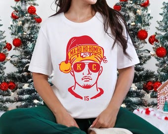 May include: White t-shirt with a red and yellow graphic of a person wearing a Santa hat and sunglasses. The hat reads "MA-HO HO-HOMES KC". The number "15" is below the graphic. The person is wearing green sweatpants.