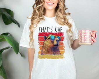 May include: White t-shirt with a graphic of a groundhog wearing sunglasses and the text "THAT'S CAP" in a retro style.