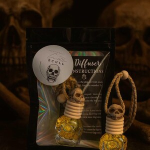 Pode incluir: Dois ambientadores de carro em forma de caveira com um frasco de vidro transparente cheio de líquido amarelo. Os ambientadores têm uma base de madeira e um cordão trançado. Uma bolsa preta com um adesivo redondo que diz "SCULL" e um gráfico de caveira está atrás dos ambientadores.