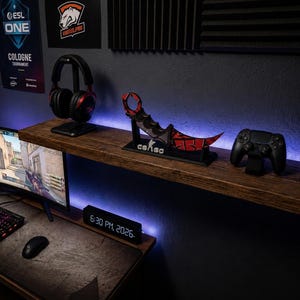 May include: A gaming setup featuring a curved monitor, a backlit keyboard, a mouse, and a digital clock displaying 6:30 PM, 2026. On a wooden shelf, there is a black and red headset, a red and black karambit knife replica with a CS:GO stand, and a controller.