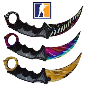 Op de afbeelding: Drie zwarte karambit-messen met verschillende gekleurde ontwerpen. Het bovenste mes heeft een zwart-wit zebra-achtig patroon. Het middelste mes heeft een regenboogpatroon. Het onderste mes heeft een houtnerfpatroon. Elk mes heeft de woorden "Counter Strike" aan de zijkant gedrukt.