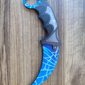 Könnte beinhalten: Ein blaues und schwarzes Karambit-Messer mit einem Spinnennetz-Design und den Worten "Counter Strike" auf dem Griff.