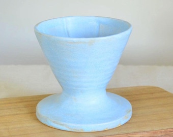 Könnte beinhalten: Ein hellblauer Keramik-Kaffee-Pour-Over-Kegel mit breitem Boden und ausgestellten Seiten. Der Pour-Over befindet sich auf einem Holzschneidebrett mit Griff. Der Kegel ist etwa 13 cm hoch.