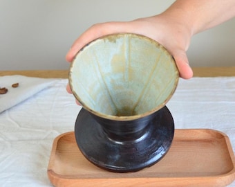 Könnte beinhalten: Ein Keramik-Pour-Over-Kaffeebereiter mit dunkelbraunem Sockel und hellblauem Innenraum. Der Kaffeebereiter steht auf einem kleinen Holztablett. Kaffeebohnen sind auf einem weißen Tuch im Hintergrund verstreut. Der Kaffeebereiter wird von einer Person gehalten.