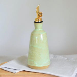 May include: A light green ceramic oil bottle with a gold spout. The bottle has a rounded base and a slender neck. It sits on a wooden cutting board with a white cloth. The bottle is approximately 20 cm tall.