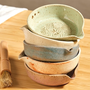 Può includere: Una pila di cinque piccoli grattugi per l'aglio in ceramica in vari colori: verde chiaro, beige, blu, arancione e rosa. Ogni grattugia ha un beccuccio e una superficie di grattugia strutturata. Sono visibili anche un piccolo pennello e spicchi d'aglio.