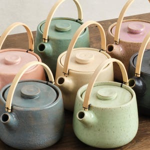 以下が含まれることがあります： ピンク、グリーン、ブルーなど、さまざまなパステルカラーの小さなセラミックティーポットのコレクション。各ティーポットには、お揃いの蓋と明るい色の木製ハンドルが付いています。ティーポットは木製の表面に配置されています。