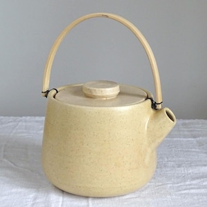 Può includere: Teiera in ceramica beige con manico e coperchio in bambù. La teiera ha un corpo arrotondato e un beccuccio. Il manico è fissato con filo nero. La teiera è su una superficie bianca.