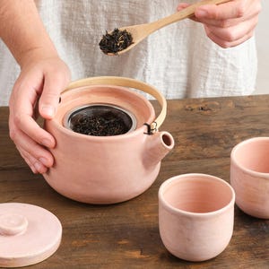 Peut inclure: Une théière en céramique rose avec une anse en bambou et un couvercle assorti, ainsi que deux petites tasses. Une cuillère en bois contient des feuilles de thé en vrac, prêtes à être ajoutées à la théière.