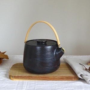 Op de afbeelding: Een donkerblauwe keramische theepot met een lichtgekleurde houten handgreep. De theepot staat op een houten snijplank. Een witte doek en gedroogde bladeren zijn op de achtergrond.
