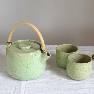 Może przedstawiać: Jasnozielony ceramiczny imbryk z bambusowym uchwytem i dwoma pasującymi filiżankami. Imbryk i filiżanki mają nakrapiany wzór. Zestaw stoi na białym obrusie, sugerując zestaw do herbaty.