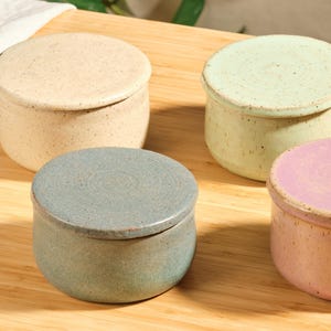 Peut inclure: Quatre petits pots ronds en céramique avec couvercles de couleurs pastel : beige, vert, bleu-gris et rose. Les pots sont posés sur une surface en bois, avec un feuillage vert en arrière-plan. Les pots ont une texture mouchetée.
