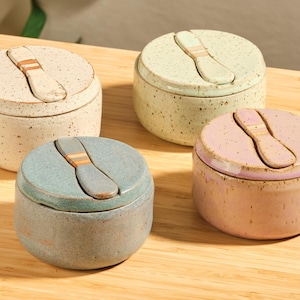 Peut inclure: Quatre petits pots ronds en céramique aux couleurs pastel : crème, vert, bleu et rose. Chaque pot a un couvercle assorti avec une poignée en forme de cuillère. Les pots sont mouchetés et reposent sur une surface en bois.