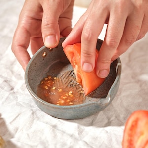 May include: A person is using a small, blue-grey ceramic tomato grater to prepare a tomato. A halved red tomato sits nearby. A wooden-handled brush with natural bristles is on the left. The scene is set on a white, textured surface.