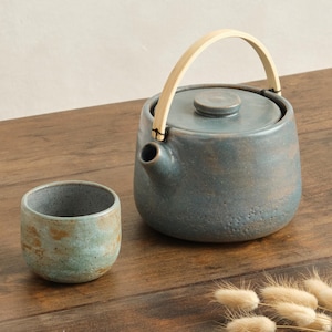 May include: A ceramic tea set with a blue-gray glaze. The set includes a teapot with a wooden handle and a small cup. The teapot is approximately 6 inches tall, and the cup is about 3 inches in diameter. The set is on a wooden surface.