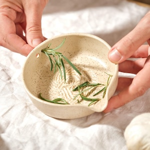 Peut inclure: Un bol râpe-ail en céramique beige moucheté avec une petite poignée, rempli de brins de romarin frais. Une brosse en bois avec des poils est posée à gauche. Le bol est tenu par des mains sur un tissu blanc.