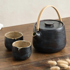 Może przedstawiać: Czarny ceramiczny czajnik z jasnym drewnianym uchwytem i pokrywką, w towarzystwie dwóch pasujących filiżanek. Czajnik ma dzióbek i stoi na drewnianej powierzchni. Filiżanki i czajnik mają nakrapiane wnętrze.