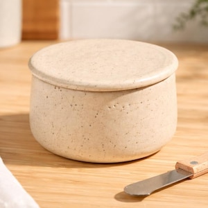 May include: A beige ceramic butter dish with a matching lid. A stainless steel butter knife with a wooden handle rests on the wooden surface. The dish is round and has a speckled texture. Kitchenware.