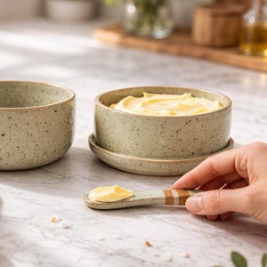 Può includere: Due burriere in ceramica maculata, una riempita di burro giallo e un coltello da burro abbinato. I piatti sono verde chiaro con macchie marroni. Il coltello da burro ha un manico verde chiaro con strisce marroni e bianche.