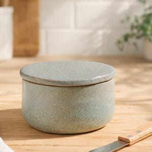 Peut inclure: Un beurrier rond en céramique bleu clair avec un couvercle assorti. Un couteau à beurre en acier inoxydable avec un manche en bois repose sur une surface en bois. Ustensiles de cuisine et planche à découper en arrière-plan.