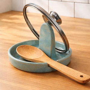 May include: A light blue ceramic spoon rest with a built-in lid holder. A wooden spoon rests in the dish. The lid holder is made of metal. The spoon rest is on a wooden surface.