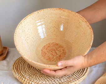 Może przedstawiać: Duża, nakrapiana ceramiczna miska z jasnobeżowym szkliwem i pomarańczowym geometrycznym wzorem na dnie. Miskę trzyma się nad plecioną podkładką. Widoczna jest również pasująca butelka na olej i drewniane łyżki.