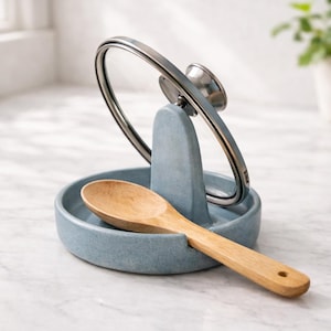 May include: A blue ceramic kitchen utensil rest with a wooden spoon and a stainless steel lid holder. The spoon is light brown. The lid holder is holding a clear glass lid with a stainless steel rim.