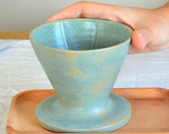 Könnte beinhalten: Ein türkisfarbener Keramik-Kaffee-Pour-Over mit einem Holztablett. Der konische Kaffeebereiter steht auf einem kleinen, rechteckigen Holztablett. Kaffeebohnen sind auf einem weißen Tuch verstreut.