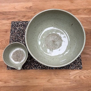 May include: Two handmade ceramic bowls in a speckled, pale green glaze. A large bowl and a smaller bowl with a spout are displayed on a dark, textured mat. The bowls have a geometric pattern in the centre.