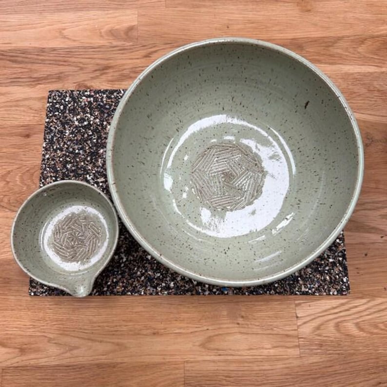 May include: Two handmade ceramic bowls in a speckled, pale green glaze. A large bowl and a smaller bowl with a spout are displayed on a dark, textured mat. The bowls have a geometric pattern in the centre.