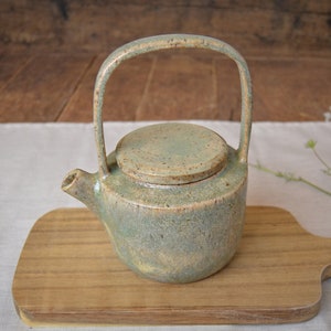 Può includere: Una teiera in ceramica verde e marrone con un coperchio e un manico. La teiera è appoggiata su un tagliere di legno.