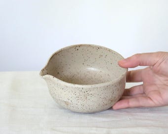 May include: A speckled, off-white ceramic bowl with a pouring spout. The handmade bowl has a rounded shape and is held by a hand. The bowl is likely used for mixing or serving food.