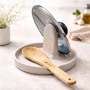 May include: A light grey ceramic spoon rest with a built-in holder for a pot lid. A wooden spoon rests in the tray. The lid is glass with a silver rim and black handle. Kitchenware.