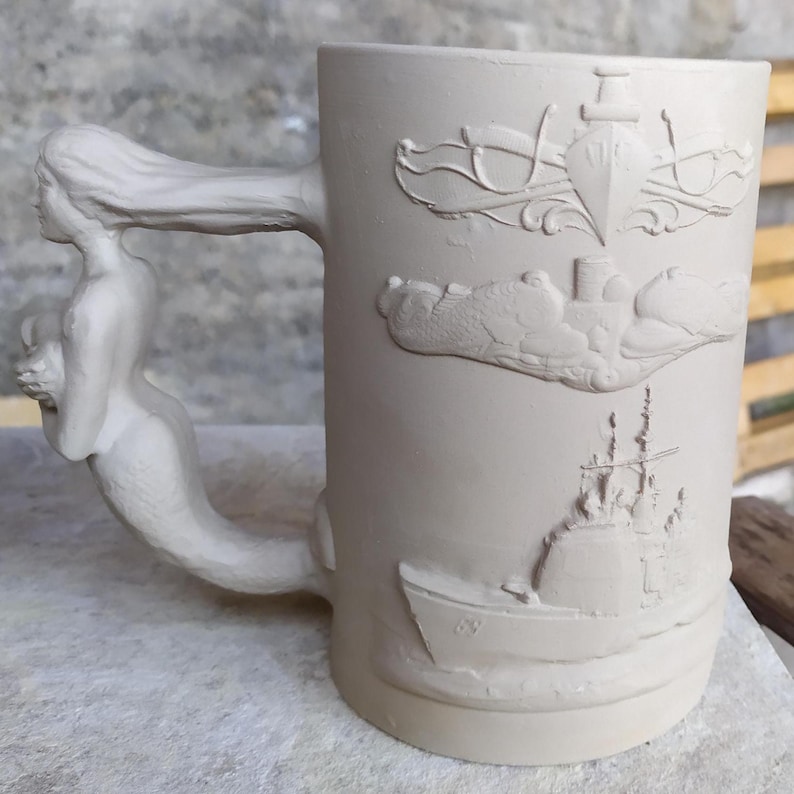 May include: A beige ceramic mug with a mermaid handle. The mug has embossed nautical designs, including a ship, a submarine, and a lighthouse. The handle is a sculpted mermaid with flowing hair.
