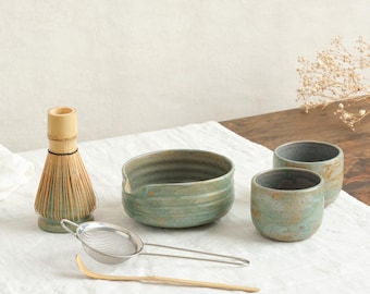 May include: A matcha tea set featuring a bamboo whisk, a ceramic bowl, two small cups, a metal sieve, and a bamboo scoop. The ceramic pieces have a mottled green and brown glaze. The whisk is light brown.