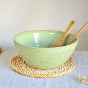 Puede incluir: Un cuenco grande de cerámica verde claro con cucharas de madera dentro, sobre un salvamanteles de paja tejida. Una botella de aceite verde claro y una pequeña bandeja de madera son visibles en el fondo.