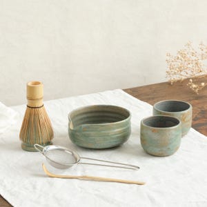 Peut inclure: Un ensemble à thé matcha comprenant un fouet en bambou, un bol en céramique, deux petites tasses, un tamis en métal et une cuillère en bambou. Les pièces en céramique ont un glaçage vert et marron moucheté.