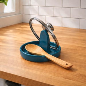 Könnte beinhalten: Ein türkisfarbener Keramik-Löffelhalter mit integriertem Deckelhalter und einem Holzlöffel. Der Löffelhalter hat eine kreisförmige Basis und einen gebogenen Metallgriffhalter. Der Holzlöffel liegt in der Basis.