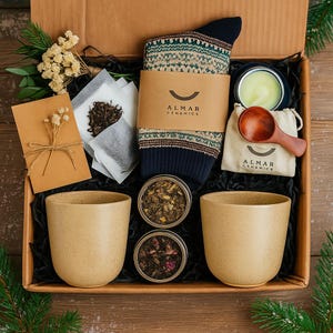 Puede incluir: Una caja de regalo llena de artículos relacionados con el té. Incluye dos tazas de cerámica beige, un par de calcetines estampados, dos latas de té, bolsitas de té, una vela pequeña, una cuchara de madera y una bolsa con "ALMAR CERAMICS".