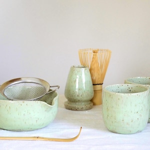 May include: A green speckled ceramic matcha bowl, whisk, and two cups. The bowl has a spout and a strainer on top. The whisk is made of bamboo. The cups are also speckled green.