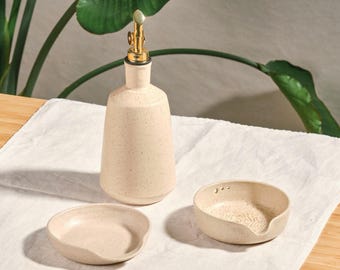 Puede incluir: Una botella de aceite de cerámica beige con boquilla dorada, acompañada de dos pequeños cuencos a juego. Los artículos están dispuestos sobre un paño blanco, con una planta verde de fondo. La botella mide unos 20 cm de altura.